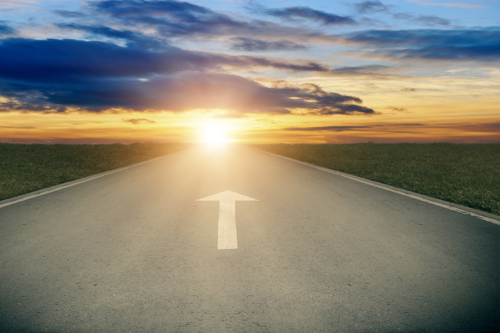 Asphalt road with white arrow sign goes to horizon. Sunshine on horizon and the road strigt away forward. Moving forward concept. Clear and positive perspectives ahead concept