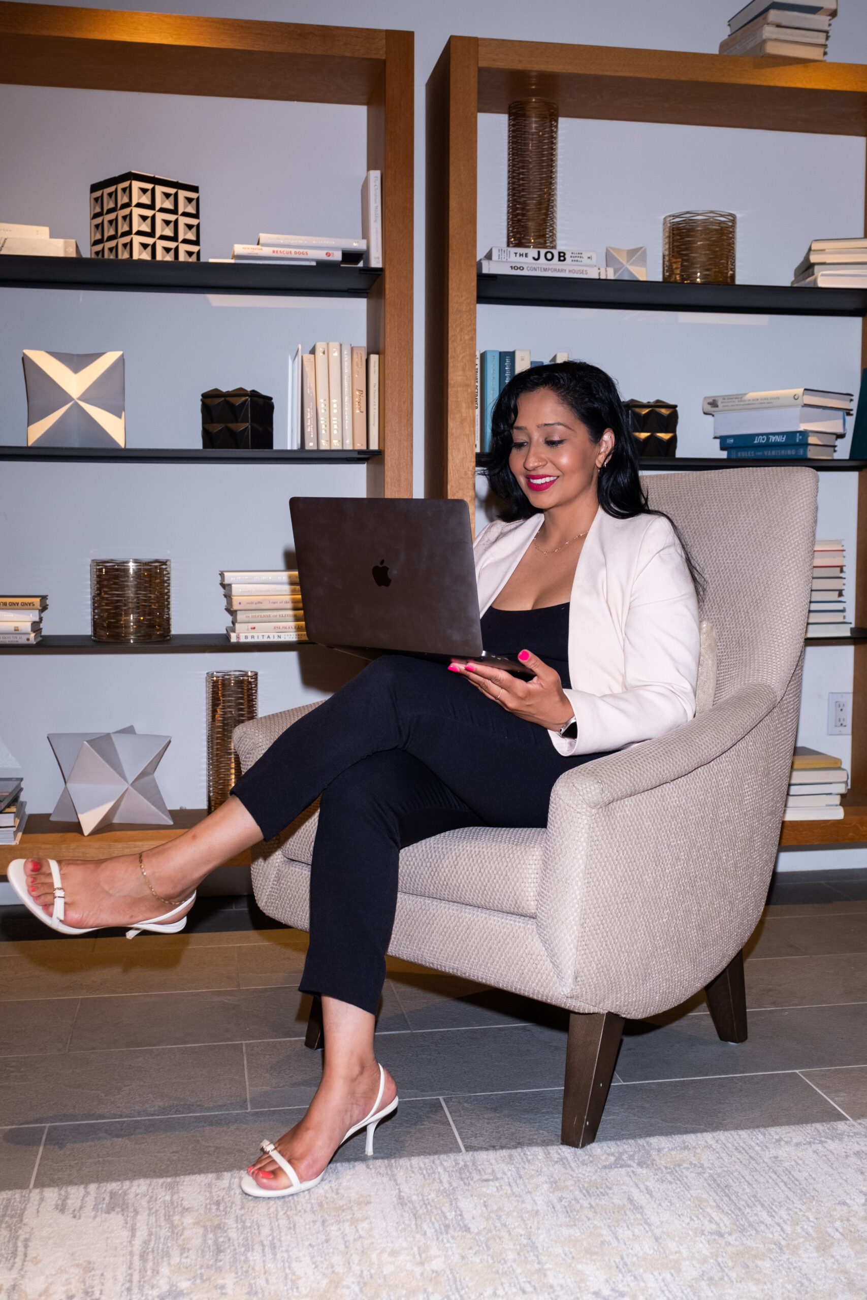 Professional woman in white blazer and black outfit sitting in modern office chair with laptop and phone, smiling while working in front of organized bookshelf