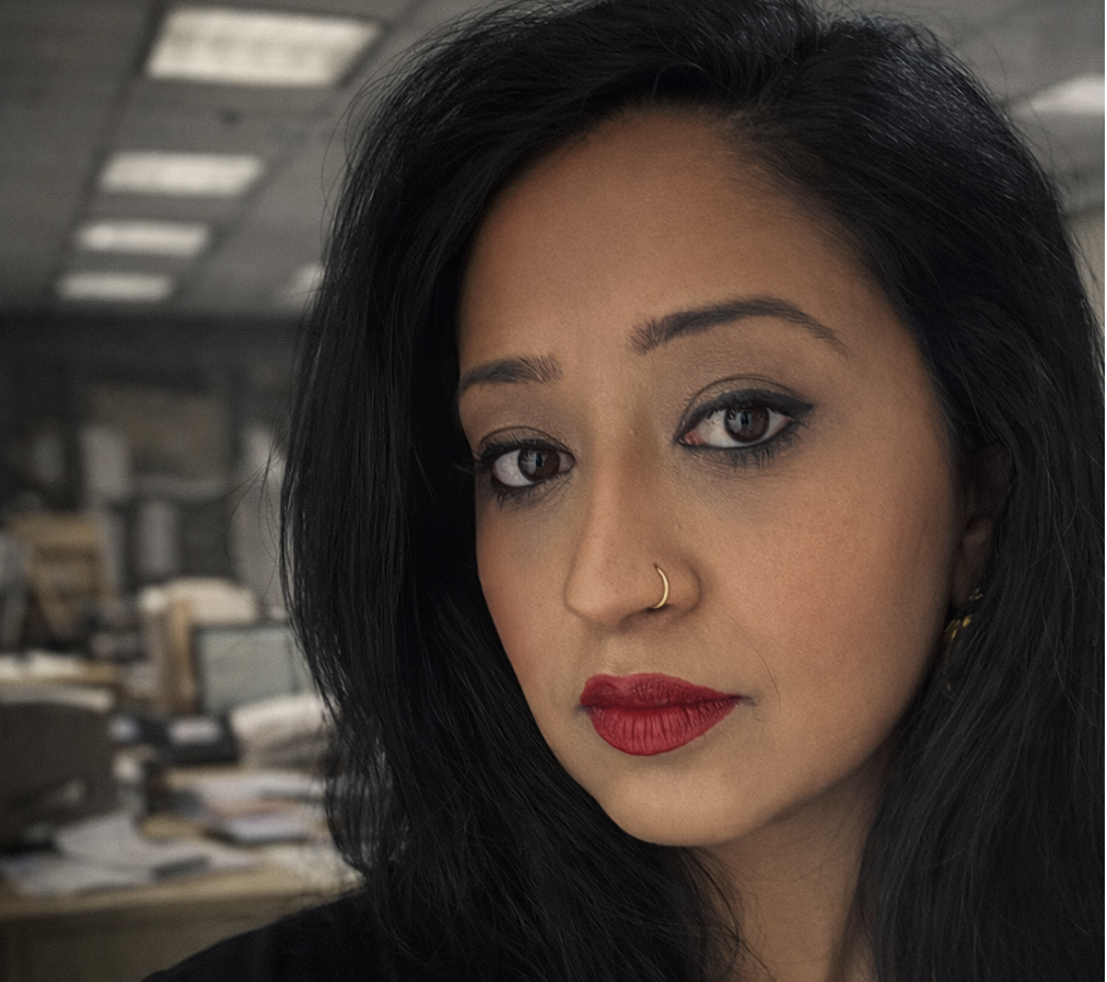 Close-up portrait of a professional woman with long dark hair and red lipstick, looking directly at the camera with a serious expression in a blurred office setting, with text overlay reading “Your worst job might be the best thing that ever happened.”